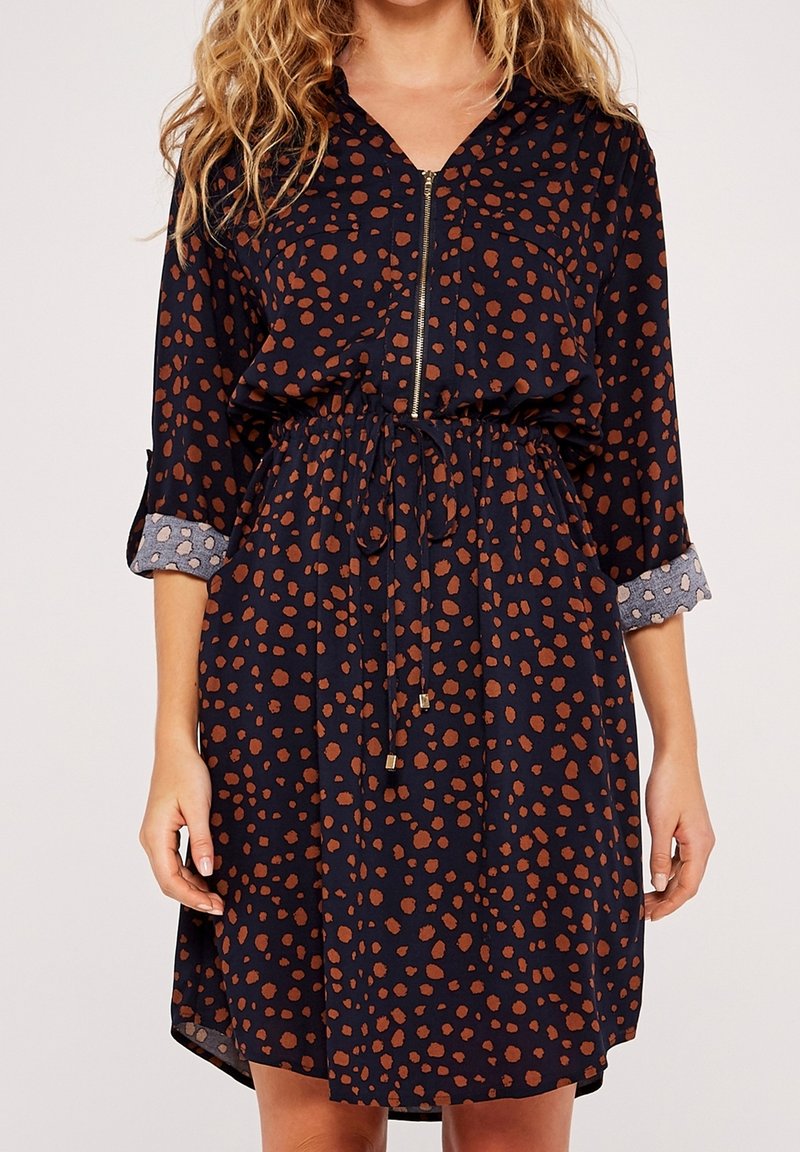 Femme portant une robe noire longueur genoux avec des taches marron, des poignets retroussés gris à motif, une fermeture éclair à l'avant et une taille à cordon.