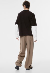 Übergroßes schwarzes Baumwoll-T-Shirt, das über ein langärmliges weißes Shirt getragen wird, kombiniert mit lockeren beigefarbenen Hosen. Enthält Seitentasche, weiche Textur und lässige Passform.