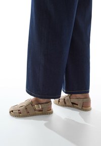 Sandalias beige con tiras entrecruzadas y diseño de punta cerrada. Presentan una hebilla en el lateral y una suela de goma texturizada. Combinadas con pantalones de mezclilla oscura de pierna ancha.