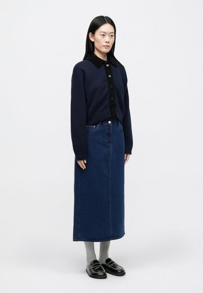 Ναυτικό πουλόβ�ερ με μαύρο γιακά από κοτλέ, κούμπωμα μπροστά με κουμπιά· μακριά μπλε τζιν φούστα· γκρίζες κάλτσες· μαύρα μοκασίνια με διακοσμητική ραφή.