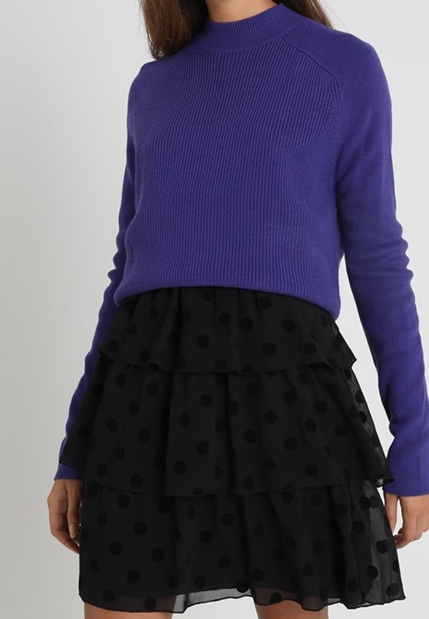 Femme portant un pull en maille violet à manches longues et une jupe à volants noire à gros pois, debout devant un fond uni.