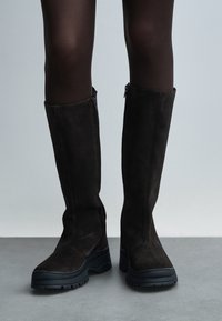 Rudos spalvos veliūriniai ilgi batai iki kelių su tekstūruotu paviršiumi, šoninėmis užtrauktukais ir stambiomis juodomis gumos padomis. Lygus viršus ir struktūruotas batraiščio vamzdelis.