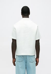 Košeľa s krátkym rukávom a golierom z ľahkej šedo-sivej textúrovanej látky, nosená s svetlo-modrými rifľami, ukazujúca pohodlný strih a jednoduchý dizajn.