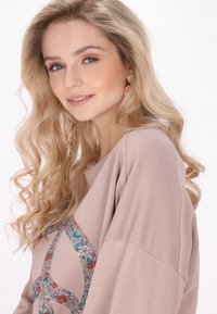 Felpa oversized rosa chiaro con un design di un segno di pace floreale. La modella ha i capelli lunghi e mossi e indossa orecchini decorativi.
