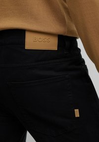 Pantalones negros con una etiqueta de cuero color crema en la parte trasera de la cintura que dice "BOSS", con costuras dobles y bolsillos traseros estándar.