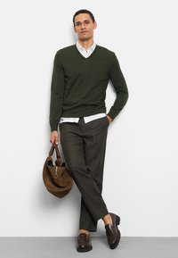 Maglione verde scuro a scollo a V sopra una camicia bianca con colletto, abbinato a pantaloni scuri e mocassini marroni, con una borsa in suede marrone in mano.