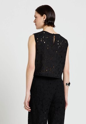 Donna con capelli corti e scuri che indossa una maglia senza maniche in pizzo floreale nero e pantaloni abbinati, vista da dietro su uno sfondo neutro.