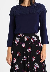 Femme portant un haut à manches longues bleu marine avec un détail en dentelle sur les épaules et une jupe noire à motif floral rouge, blanc et violet.