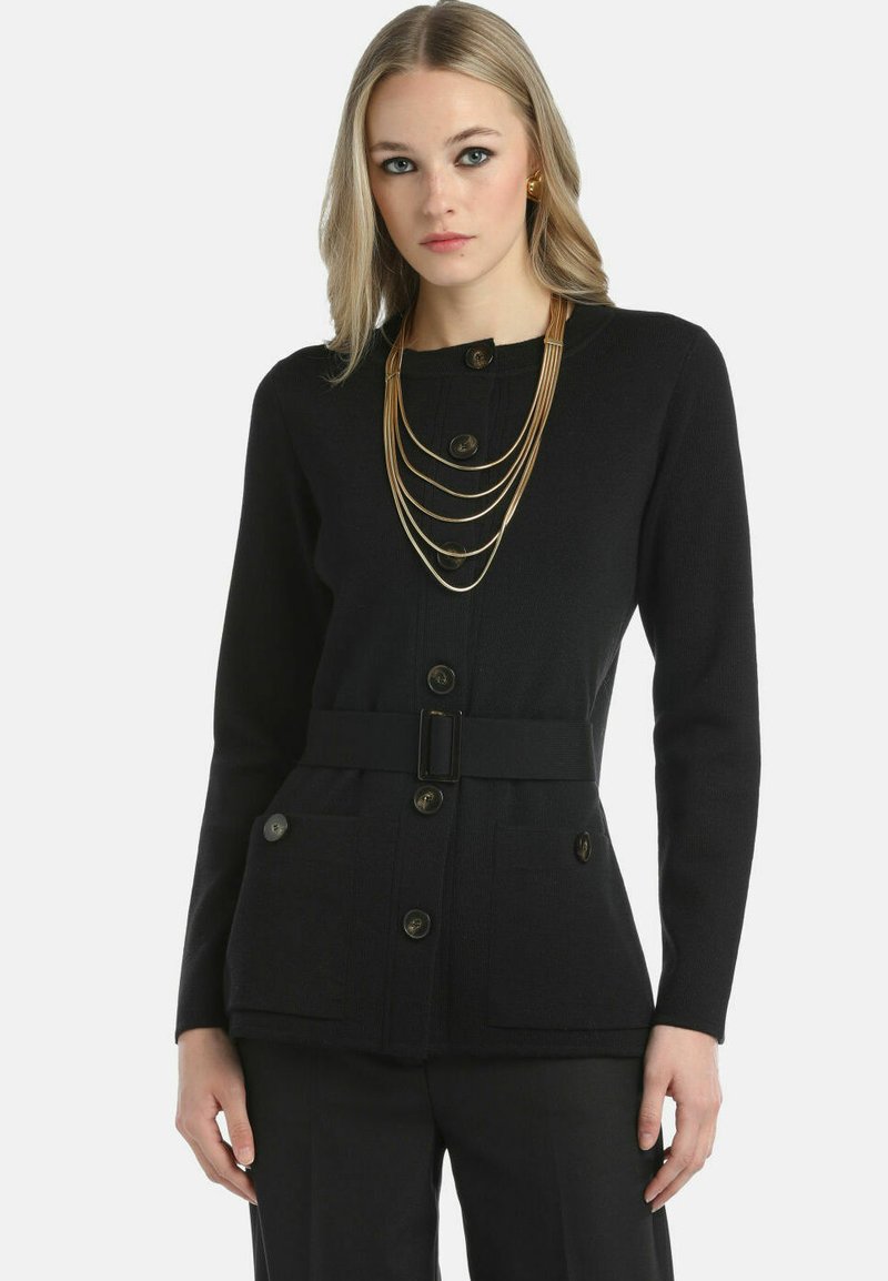 Mujer vistiendo un cárdigan negro con botones y un cinturón ancho, pantalones negros, collares de oro en capas y pendientes de oro contra un fondo simple.