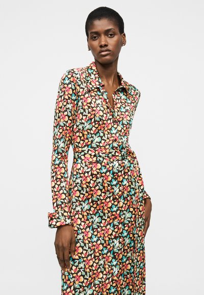 Γυναίκα που φοράει μακρυμάνικο, φλοράλ, φόρεμα τύπου wrap με γιακά, στέκεται μπροστά σε ένα απλό φωτεινό υπόβαθρο.