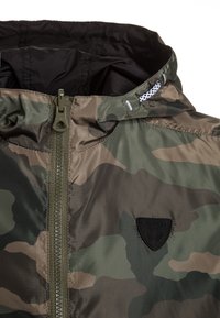 Camouflage zip-up jacket with a black lining. Features a high collar, adjustable hood, and round black logo patch on the chest.