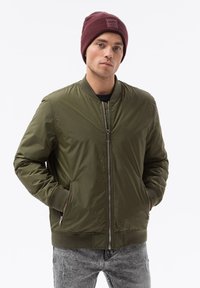 Giacca bomber verde oliva con zip frontale, polsini e colletto a coste; abbinata a un berretto bordeaux. Tessuto strutturato e vestibilità casual.