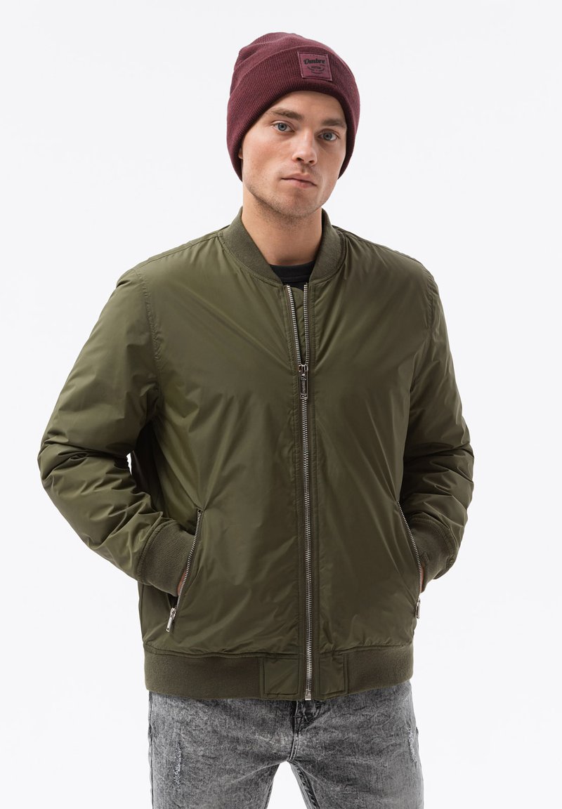 Giacca bomber verde oliva con zip frontale, polsini e colletto a coste; abbinata a un berretto bordeaux. Tessuto strutturato e vestibilità casual.