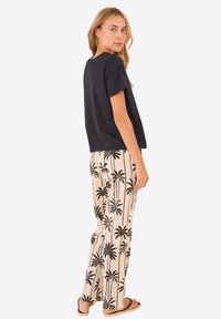 Zwarte top met korte mouwen gecombineerd met beige broek met zwarte palmbomenprints. Het model draagt zwarte sandalen en presenteert een casual outfit.