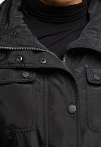 Veste légère noire avec un col montant, une fermeture éclair frontale et des boutons-pression. Comprend deux poches avant à rabats et des détails cousus.