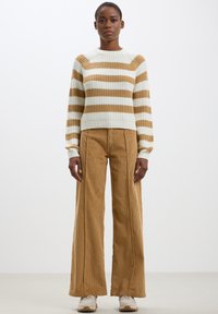 Maglione in maglia con strisce dorate e bianche; texture a coste. Abbinato a pantaloni in corduroy a gamba larga di colore beige. Ai piedi, sneaker beige.