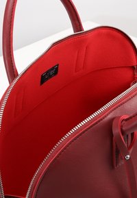 Červená kabelka s texturovaným povrchem, se zipovým otvorem a hladkým červeným interiérem. Obsahuje štítek a dvě vnitřní kapsy pro organizaci.