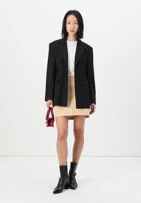 Blazer nero con spalle imbottite, mini gonna in suede beige, T-shirt bianca e stivaletti neri a punta; tiene in mano una piccola borsa rossa.