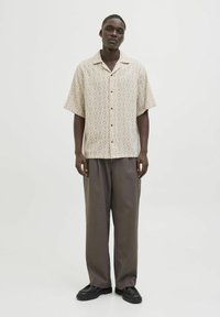 Kurzärmliges Button-Up-Hemd in Creme mit diamantförmigem Muster, kombiniert mit locker sitzenden grauen Hosen und dunklen, strukturierten Schuhen.