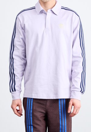 Mężczyzna ubrany w jasnofioletową koszulkę polo Adidas z długim rękawem, z granatowymi paskami na rękawach oraz ciemne spodnie z niebieskimi pionowymi paskami.