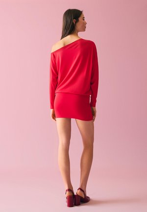 Woman with long dark hair stands facing away, wearing a red off-shoulder dress and matching heels against a pink background.