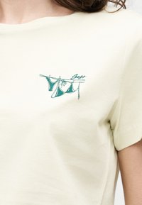 Μπεζ μπλουζάκι με μικρή τυρκουάζ κεντητική με κομμάτια μπικίνι κρεμασμένα σε μία απλώστρα στην άνω περιοχή του στήθους.