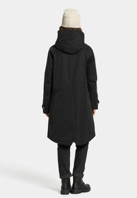 Zwarte waterdichte parka met een capuchon, lengte tot halverwege de dij en een gebogen onderkant. Getextureerde stof en met knopen sluitende manchetten. Gecombineerd met zwarte broeken en laarzen.