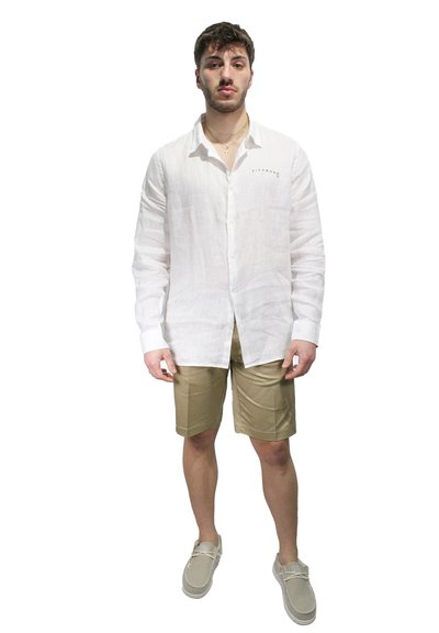 Camicia bianca in lino a maniche lunghe con chiusura a bottoni, abbinata a pantaloni beige realizzati in materiale liscio. Accessori includono un piccolo logo e scarpe informali.