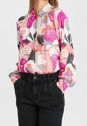 Femme portant un chemisier à manches longues boutonné avec un motif floral rose, orange et gris rentré dans un pantalon noir taille haute, les mains dans les poches.