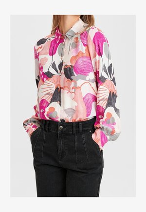 Kvinde iført en langærmet knap-op bluse med pink, orange og gråt blomsterprint, stoppet ned i højtaljede sorte bukser med hænderne i lommerne.