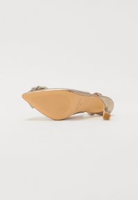 Zapato de tacón alto beige con un acabado brillante, que presenta un acento de perla y una suela texturizada. Tacón cuadrado y diseño de tiras abiertas.
