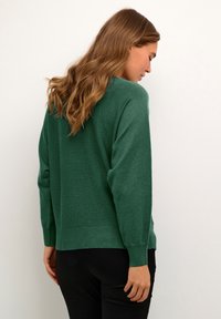 Roheline kudum, mis on lõdva lõikega, omab pikki varrukasid ja soonikuid. Sile tekstuur ja lihtne disain.