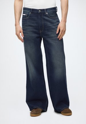 Mand iført vide mørkeblå jeans og brune sko, stående med hænderne afslappet langs siden mod en ensfarvet baggrund.