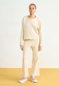 Beige pullover en bijpassende wijde broek van gestructureerde stof. Model draagt roze platform sneakers. Achtergrond heeft een lichte, neutrale kleur.