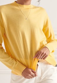 Person som bär en ljusgul långärmad tröja, justerar nederkanten, tillsammans med off-white byxor och ett silversmycke med en halvmånependel.