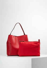 Červená kožená taška přes rameno s hladkým povrchem, se boční kapsou a krátkým držadlem, vedle níž se nachází malá červená zipová taštička.