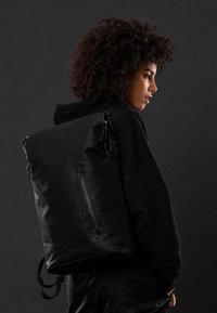 Zaino a tracolla nero con finitura strutturata, tasca laterale con zip e spallacci regolabili. Presenta un logo discreto sul davanti.