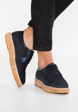 Chaussures oxford en cuir noir avec semelles plateformes en liège portées avec un jean noir, montrées sur des pieds croisés sur fond blanc.