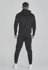 Crna dukserica s kapuljačom i odgovarajuće joggers hlače izrađene od mekanog materijala, s kapuljačom i rebrastim manžetama. Uključuje minimalistički dizajn.