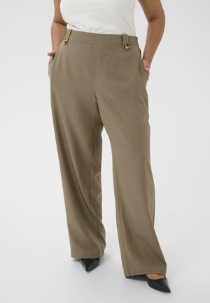 Femme portant un pantalon kaki à jambes larges avec des boutons à l'avant et des chaussures noires à bouts pointus, les mains dans les poches, debout devant un fond clair.