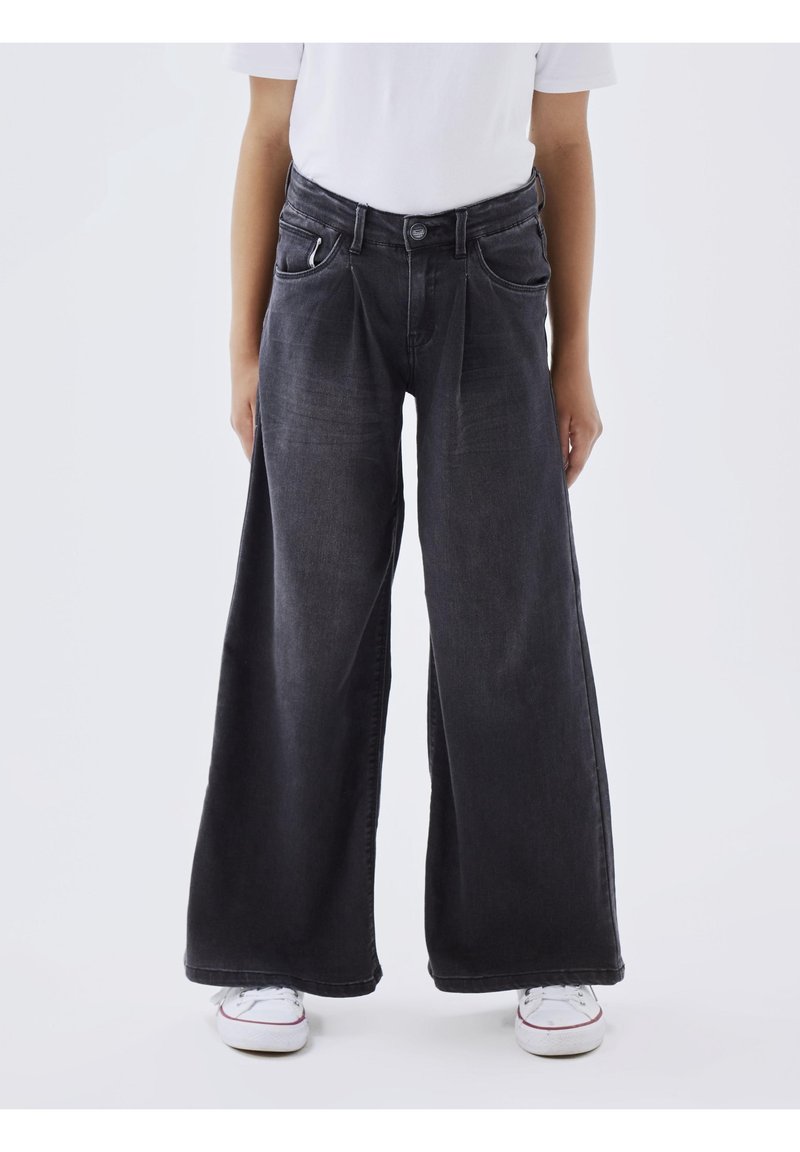 Personne portant un jean noir taille haute à jambes larges, un t-shirt blanc et des baskets blanches, debout devant un fond clair uni.