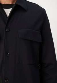 Primo piano di una camicia nera con bottoni, dotata di una grande tasca a patta sul petto, indossata sopra un top bianco e nero.