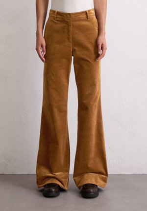 Persona que lleva pantalones de pana marrones de talle alto y pierna ancha y zapatos negros, de pie frente a una pared lisa y clara.