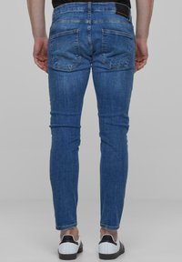 Slim-fit siniset farkut, joissa on haalistunut pinta, viisi taskua ja musta etiketti vyötäröllä, yhdistettynä valkoisiin kenkiin.