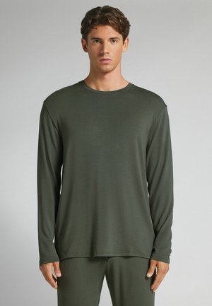 Junger Mann, der ein schlichtes dunkelgrünes Langarmshirt und passende Hosen trägt, steht frontal vor einem neutralen Hintergrund.