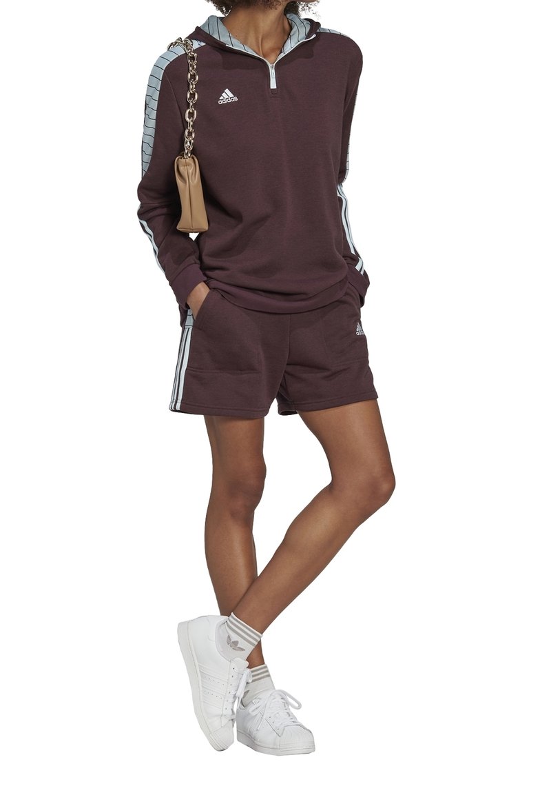 Personne portant un sweat à capuche Adidas bordeaux foncé et un short assorti avec des bandes bleu clair, des chaussettes blanches, des baskets blanches et un sac beige.