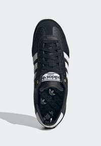 Black sneaker with perforated texture and white three stripes. Features a black lacing system, gold accents, and a patterned insole.