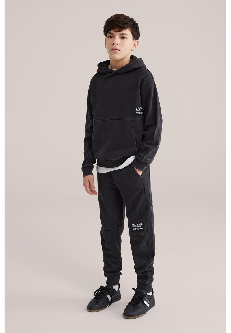 Teismeline poiss seisab, seljas must kapuutsiga sviiter ja sobivad dressipüksid, millel on kiri "SALTY DOG", mustad tossud ja valged sokid heledal taustal.