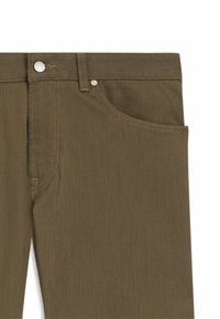 Primo piano di pantaloni di denim verde oliva che mostrano la tasca anteriore, la passante per cintura, i dettagli delle cuciture e il bottone e rivetto in argento.