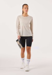 Beige sporttop met lange mouwen, zwarte fietsbroek, doorzichtige waterfles met zwarte dop, witte sokken met logo en witte sneakers.